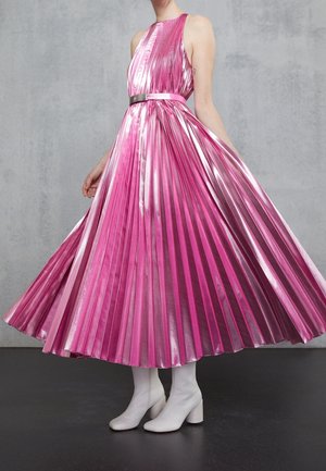 Kobieta w błyszczącej różowej plisowanej sukience midi z paskiem, w połączeniu z białymi botkami na obcasie, stojąca na szarym tle.