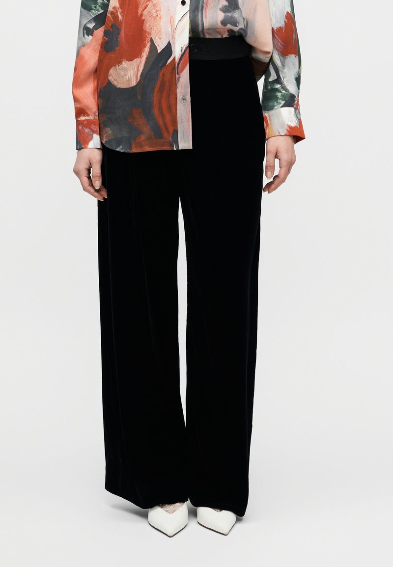 Pantaloni largi din catifea neagră cu o textură netedă, asortați cu o bluză colorată cu imprimeu floral în nuanțe de roșu, verde și gri.