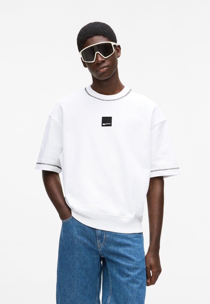 Fehér, túlságosan nagy, rövid ujjú pulóver fekete logóval és szürke részletekkel a gallérnál és a mandzsettánál. Kék farmernadrág és nagy napszemüveg.