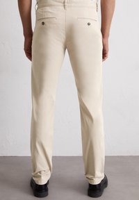 Beige katoenen broeken met een rechte pijp, met twee achterzakken met metalen hardware en een gladde textuur. Draagt zwarte schoenen.