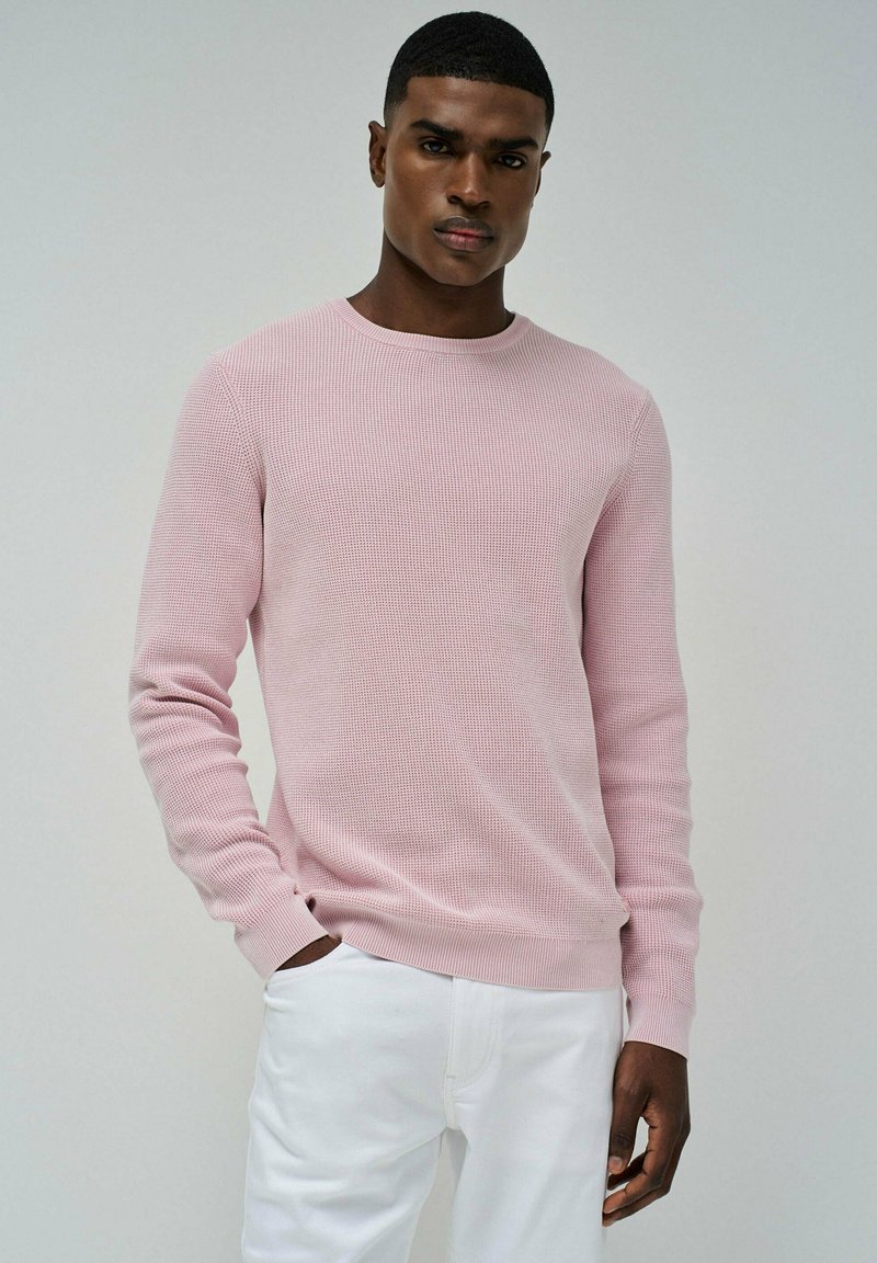 Maglione a coste rosa chiaro con scollatura rotonda e maniche lunghe, abbinato a pantaloni bianchi. Texture morbida, design minimale e forma aderente.