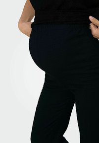 Personne enceinte portant un pantalon noir texturé et un haut noir, la main reposant sur le bas du dos et la hanche.