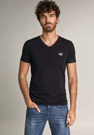 Camiseta negra de cuello en V hecha de una mezcla de algodón, con un diseño ajustado, mangas cortas, que presenta un pequeño parche de logo en el lado izquierdo del pecho, combinada con jeans azules.