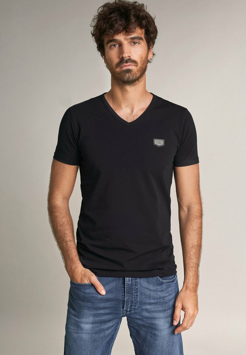 Camiseta negra de cuello en V hecha de una mezcla de algodón, con un diseño ajustado, mangas cortas, que presenta un pequeño parche de logo en el lado izquierdo del pecho, combinada con jeans azules.