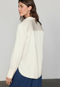 Lys beige langærmet skjorte med en kontrasterende beige rygpanel, knapmanchetter og en rund afslutning, lavet af et glat stof.