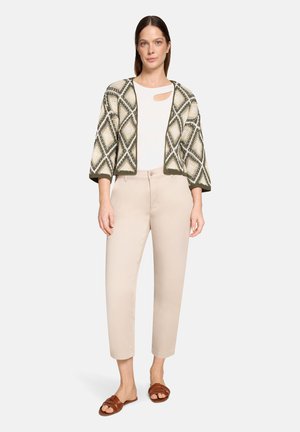 Frau trägt beige Hosen, weißes Oberteil mit ausgeschnittenen Schultern, olivgrünen Strickcardigan mit Muster und braune Slip-On-Sandalen, steht vor weißem Hintergrund.