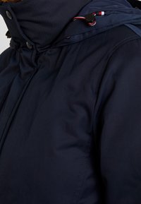 Marinblå jacka med hög krage, dragkedja och knappstängning, samt justerbar dragsko i huvan. Innehåller slätt, vattentåligt tyg.