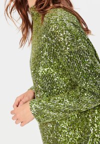 Donna che indossa un abito con maniche lunghe, verde smeraldo con paillettes e texture scintillante, mostrata da metà torace alle spalle e alle mani.