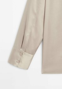 Poignet de chemise beige à manches longues avec deux boutons, tissu lisse et détails de couture visibles sur fond blanc.