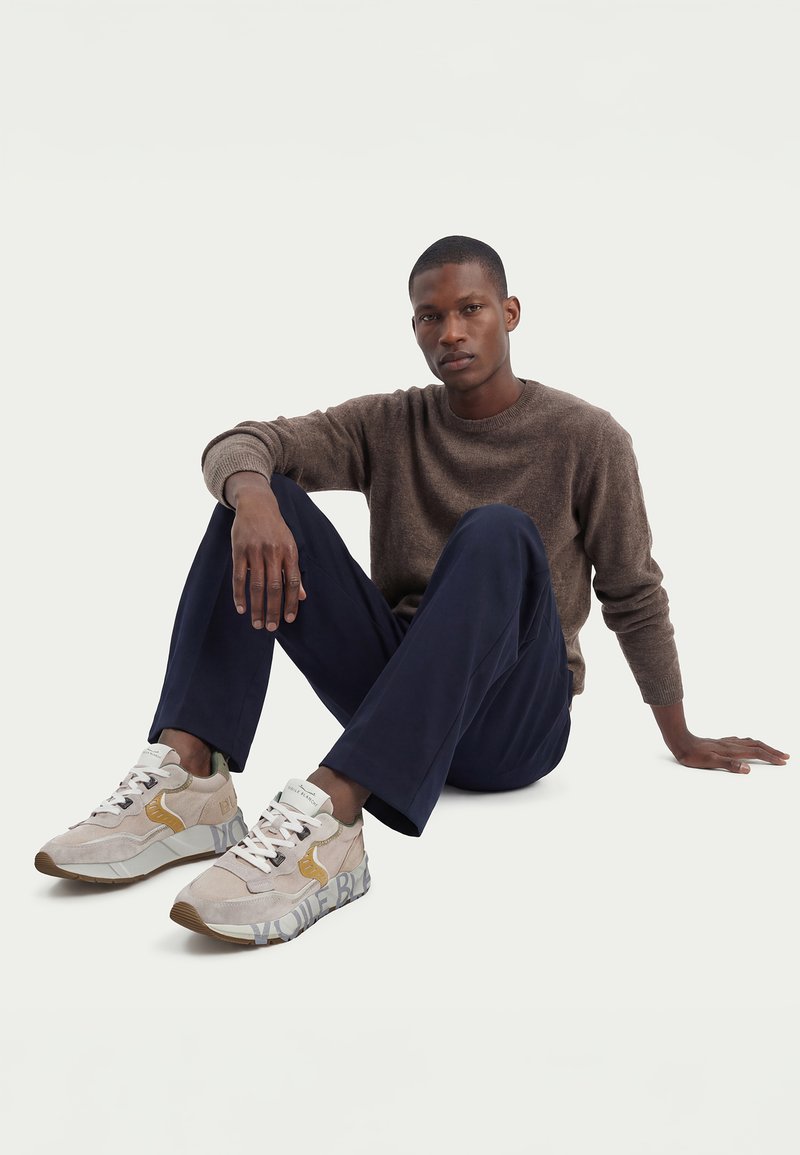 Homme assis par terre portant un pull marron, un pantalon bleu marine et des baskets beiges avec des détails jaunes sur un fond clair uni.