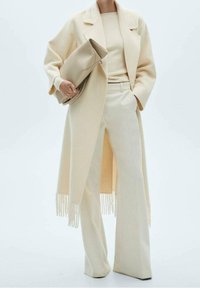 Manteau long beige à ourlet à franges, haut beige côtelé et pantalon à jambes larges, tous dans des nuances tonales assorties, tenant une grande pochette beige.