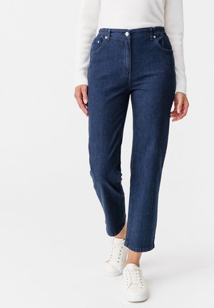 Jean droit en denim bleu foncé avec cinq poches, fermeture par bouton et zip, et taille élastiquée pour plus de confort. Sneakers blanches portées.