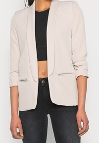 Jasno beżowa marynarka o zdefiniowanej konstrukcji, z otwartym przodem i złożonymi rękawami; ma zamykane na zamek kieszenie i gładką fakturę.