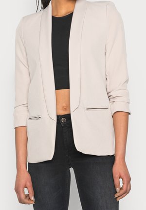 Licht beige blazer met een gestructureerd ontwerp, open front en omgeslagen mouwen; voorzien van ritszakken en een gladde textuur.