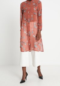 Rose-colored sheer blouse with floral pattern, button-down front, long sleeves, and single chest pocket. Paired with wide white trousers.