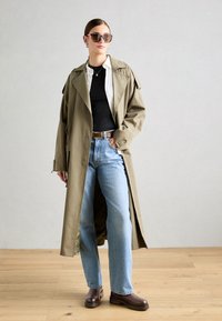Mujer vestida con un abrigo de trench beige oversized, camiseta negra, camisa blanca, jeans azul claro, botas marrones, gafas de sol y collar dorado, de pie sobre un suelo de madera.
