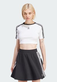 Biele skrátené tričko s čiernym golierom a logom Adidas; v kombinácii s čiernou rozšírenou sukňou s bielymi bočnými pruhmi a elastickým pásom.