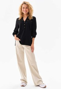 Schwarze Bluse mit langen Ärmeln, V-Ausschnitt und Knopfdetails, kombiniert mit hellbeigen, weit geschnittenen Hosen und weißen Sneakers.