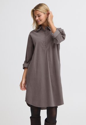 Femme portant une robe grise ample boutonnée, des collants noirs et des bottes noires, souriante et touchant ses cheveux sur un fond clair uni.