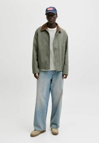 Person iført en navy kasket, olivengrøn jakke med brun krave, stribet skjorte, brede lyseblå jeans og beige sko, stående på en hvid baggrund.