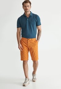 Polo-shirt bleu à manches courtes, assorti à un short orange avec des motifs noirs et des chaussures de sport gris clair.