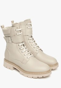 Bottines en cuir beige avec un laçage à l'avant, un col rembourré, un détail en boucle, et une semelle en caoutchouc épaisse avec une bordure contrastante en beige clair.