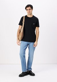 Camiseta negra de manga corta con un bolsillo, jeans de mezclilla azules y zapatos negros; modelo sosteniendo un bolso de hombro beige. Fondo liso.