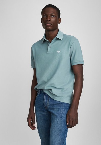 Lys grøn polo-shirt med krave og korte ærmer, forsynet med et lille broderet logo på brystet. Båret med blå jeans.
