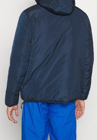 Chaqueta acolchada azul marino con capucha, que presenta una textura suave, puños elásticos y un cierre de cremallera en el dobladillo. Detalle de costura visible en la parte posterior.