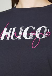 Námořnická modrá trička s kulatým výstřihem s nápisem "HUGO" v bílé a růžové barvě, strukturovaný vzor na látce.