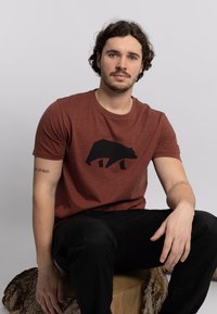 Camiseta color óxido hecha de una tela suave con un gráfico de la silueta de un oso negro en el centro. Ajuste casual con mangas cortas.