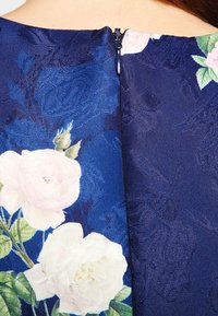 Laivastonsininen mekko, jossa on kukkakuosi valkoisilla ruusuilla. Rakennekangas, jossa on vetoketjurakenne takana.