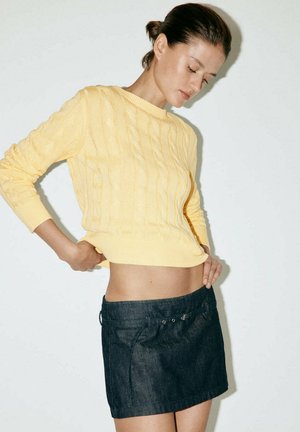Mujer con cabello oscuro recogido, lleva un jersey amarillo de punto con trenzas y una minifalda vaquera oscura, de pie frente a un fondo claro y simple.