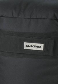Svart Dakine-väska med en slät tygtextur, vadderad topp och en rektangulär logotyp med silverdetaljer på en svart bakgrund.