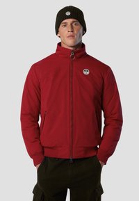 Giacca a vento rossa con collo alto, tasche con zip e patch logo. Abbinata a un berretto a coste verde scuro. Tessuto morbido, vestibilità casual.