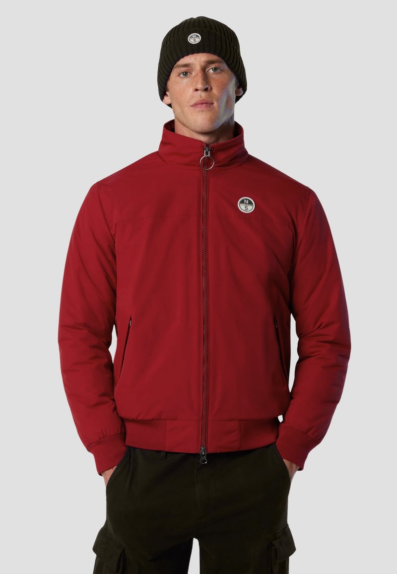 Giacca a vento rossa con collo alto, tasche con zip e patch logo. Abbinata a un berretto a coste verde scuro. Tessuto morbido, vestibilità casual.