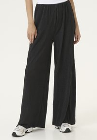 Black, pleated wide-leg pants with an elastic waistband, featuring a textured fabric. Paired with white and gray athletic shoes.