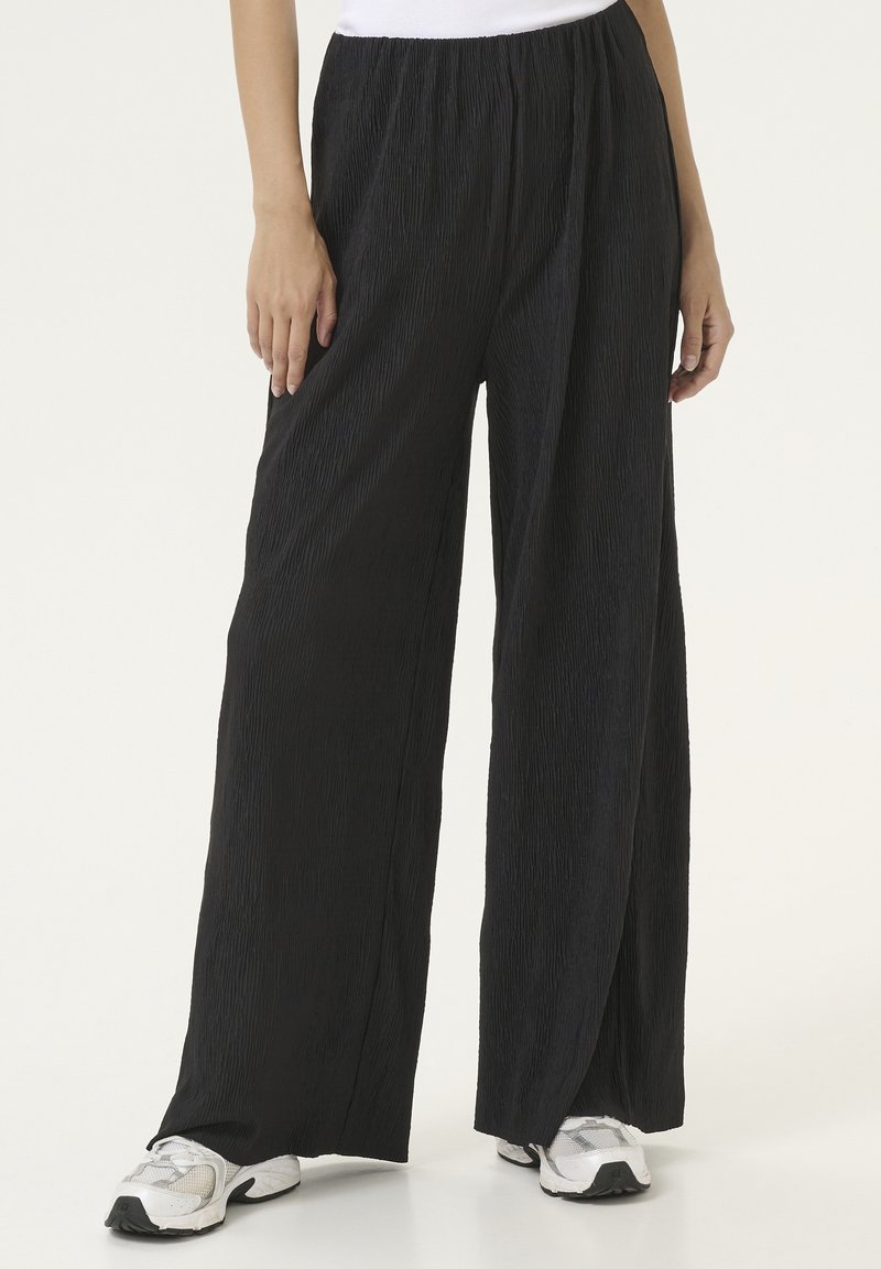 Black, pleated wide-leg pants with an elastic waistband, featuring a textured fabric. Paired with white and gray athletic shoes.