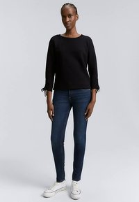 Felpa nera con maniche a tre quarti, caratterizzata da fiocchi sui polsini, abbinata a jeans skinny blu scuro e sneaker bianche.