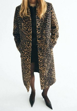 Femme portant un long manteau imprimé léopard sur une robe noire, des collants noirs transparents et des talons noirs pointus, debout les mains dans les poches.