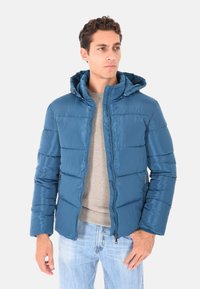 Giacca blu imbottita con finitura lucida, cappuccio e chiusura con zip. Presenta cuciture orizzontali e tasche laterali, indossata sopra un maglione grigio.