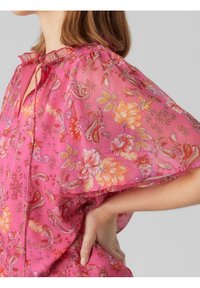 Blouse fleurie rose en tissu transparent, col à volants et manches courtes évasées. Motifs cachemire et floraux en orange et violet.