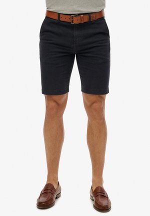 Pantalones cortos de algodón navy con un corte ajustado, que incluyen un cinturón de cuero marrón. Textura suave; dobladillo recto. Completados con zapatos marrones de slip-on.