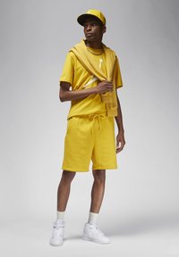 T-shirt di cotone gialla e pantaloni corti con una grafica bianca, accompagnati da un berretto giallo abbinato e un maglione drappeggiato sulle spalle. Sneakers bianche.