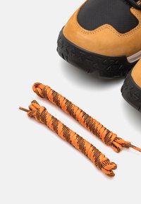 Brūnas zamšādas un melnas sieta sporta apavi ar teksturētu gumijas zoli. Ietver oranžus un melnus svītrojumus blakus apaviem.