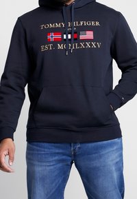 Navy hoodie made of cotton blend, featuring gold text logos and three flags. Includes a kangaroo pocket and adjustable drawstring hood.