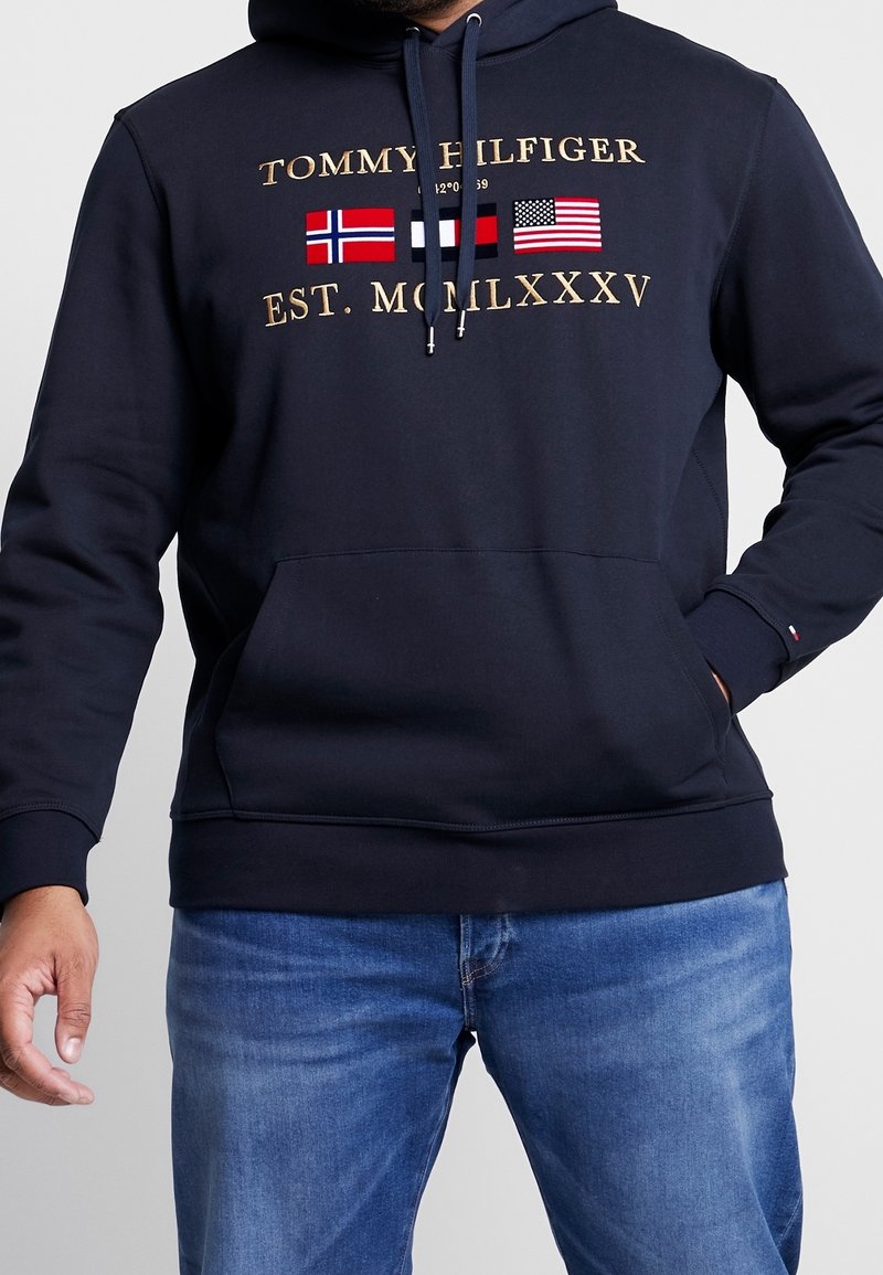 Navy hoodie made of cotton blend, featuring gold text logos and three flags. Includes a kangaroo pocket and adjustable drawstring hood.