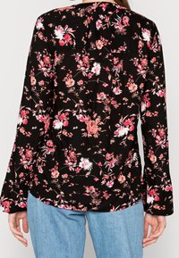 Blouse noire à motifs floraux roses et blancs, manches longues, tissu léger et ourlet arrondi. Associée à un jean bleu clair.