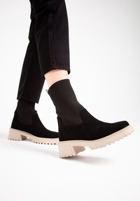 Czarne botki zamszowe z prążkowanymi elastycznymi panelami i masywną beżową podeszwą. Projekt obejmuje zaokrąglony nos i teksturowaną przyczepność na spodzie.
