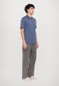 Menns kortermet polo med glidelåskrage i dempet blått stoff. Kombinert med grå vide jeans og hvite joggesko. Enkelt design, glatt tekstur.